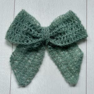 May include: A large, sage green, crocheted bow. The bow is made of a soft, fuzzy yarn and has a delicate, openwork design.