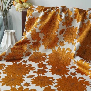 Könnte beinhalten: Weißer Stoff mit einem sich wiederholenden Muster aus großen, goldgelben Chrysanthemenblüten. Der Stoff ist drapiert und zeigt die Textur und Falten. Die Blumen haben detaillierte Umrisse und füllen die Stoffoberfläche.