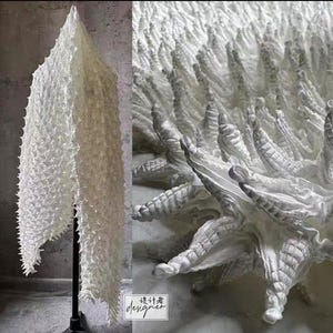 Può includere: Un'opera d'arte tessile bianca e strutturata con un design a spillo tridimensionale. L'opera è esposta su un supporto nero, che mostra i suoi intricati dettagli e la sua forma unica. L'immagine include anche un primo piano della superficie strutturata.