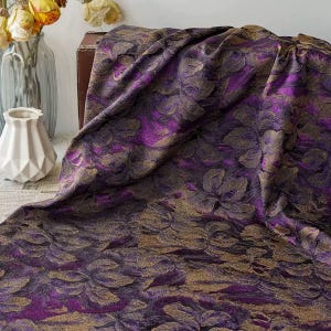 Op de afbeelding: Een luxe stof met een bloemenpatroon in paarse en gouden tinten. De stof is over een oppervlak gedrapeerd, wat de textuur en het ontwerp benadrukt. Het bloemenpatroon is gedetailleerd en voegt diepte toe aan de stof.
