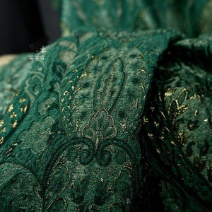 May include: Detailed shot of a dark green fabric featuring a paisley design. The material has a plush, velvety feel and is adorned with gold metallic detailing. The pattern is complex and rich in detail, creating a luxurious look.