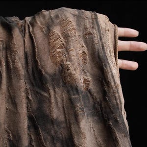 May include: Close-up of a distressed fabric in brown and black. The material features multiple tears and frayed edges, giving it a textured, worn look. The fabric is held by a hand, highlighting its drape and texture against a dark background.