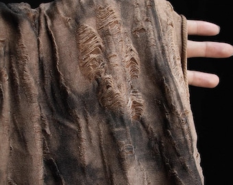 May include: Close-up of a distressed fabric with a brown and black color scheme. The fabric has numerous tears and frayed edges, creating a textured and worn appearance. The material is held by a hand, showcasing the fabric's drape.