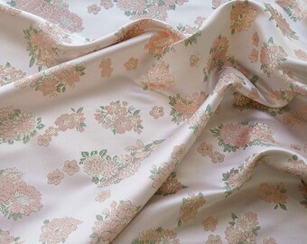 May include: A pale pink fabric with a floral pattern. The design features clusters of pink and peach flowers with green leaves and gold accents. The fabric is draped, showing the texture and sheen of the material.