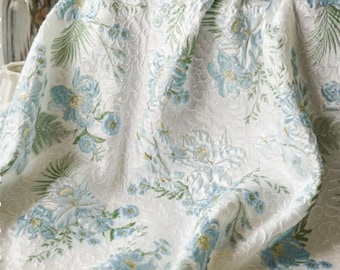 May include: A white floral patterned fabric with blue and green accents. The design features detailed flowers and foliage, creating a textured, elegant look. The fabric is draped, showcasing the intricate pattern.