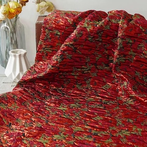 Può includere: Primo piano di un tessuto rosso strutturato con un motivo floreale in verde e nero. Il tessuto è drappeggiato, mostrando pieghe e una qualità scintillante. L'effetto complessivo è ricco e decorativo.