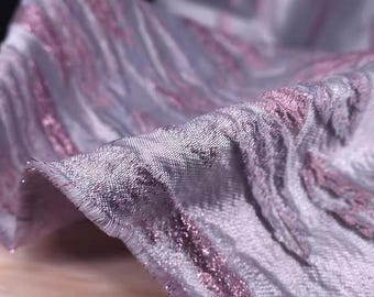 May include: Close-up of a shimmering fabric with a textured surface. The fabric is primarily a light lavender color with streaks of pink and silver running through it. The material appears to be soft and flowing, with a subtle sheen.