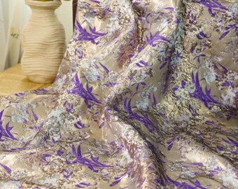 May include: A draped fabric with a metallic gold base, featuring a floral design in purple and silver. The fabric has a textured finish and a subtle sheen. A vase of flowers is visible in the background.