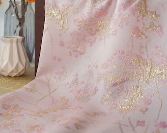 May include: A light pink fabric with a floral pattern in gold and pink. The fabric is draped, showing the texture and design. The floral pattern appears to be a stylized cherry blossom design.