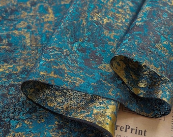 May include: Close-up of a folded fabric with a teal and gold abstract pattern. The fabric has a textured appearance with a dark background. The gold accents create a shimmering effect, adding depth and visual interest to the textile.