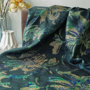 May include: Dark teal fabric with a floral pattern in gold, green, and blue. The fabric is draped, showing the texture and design. The background includes a white vase and a glass vase with yellow flowers.