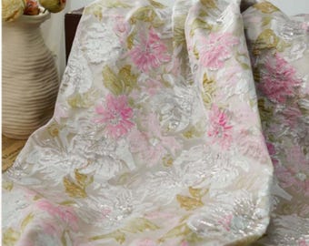 May include: Cream-colored fabric with a floral pattern featuring pink and white flowers and green leaves. The fabric has a textured, shimmering appearance, with a soft drape. The design is a repeating pattern of stylized flowers and foliage.