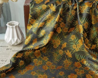 May include: Dark gray fabric with a repeating floral pattern in yellow and green. The fabric has a silky sheen and is draped over a surface. The floral design includes stylized flowers and leaves, creating a tropical or botanical theme.