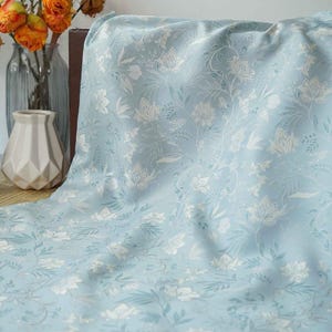 Pode incluir: Tecido azul claro com um delicado padrão floral em branco e verde claro. O tecido está drapeado, mostrando os detalhes intrincados do design. A imagem também inclui um vaso com flores.