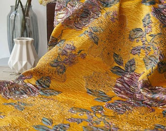 May include: A close-up of a textured, golden-yellow fabric with a floral pattern in shades of purple, blue, and gold. The fabric is draped, revealing its intricate design and sheen. The floral pattern includes large, stylized flowers and leaves.