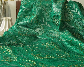 May include: Emerald green fabric with a paisley pattern and gold accents. The fabric is draped over a surface, showcasing its texture and design. The fabric's intricate details and rich color create a luxurious appearance.