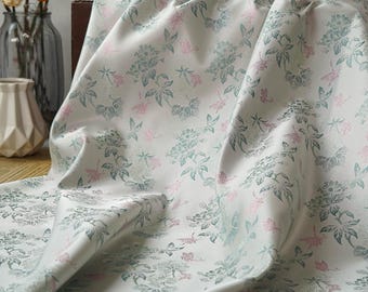 May include: Off-white fabric with a floral pattern in shades of green and pink. The fabric is draped, showing the texture and design. The flowers and leaves create a classic, elegant look, suitable for various textile projects.