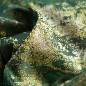 May include: Close-up of a luxurious fabric with a textured, speckled design. The material features a blend of deep green, gold, and silver hues, creating a shimmering effect. The fabric is folded, revealing its layered construction and sheen.