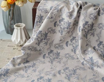 May include: Cream-colored fabric with a blue floral pattern. The fabric is draped, showing the intricate design of flowers and leaves. A vase of yellow flowers and a white geometric vase are in the background.