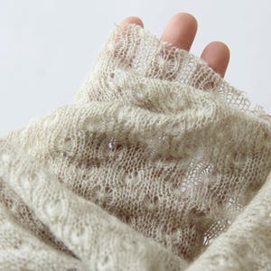 May include: Close-up of a beige, open-weave fabric. The fabric has a delicate, lacy texture with small, repeating floral patterns. The material appears soft and lightweight, ideal for a scarf or shawl.