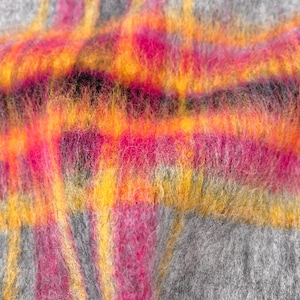 Puede incluir: Primer plano de un textil suave y texturizado con un patrón de cuadros. El diseño presenta colores vibrantes, incluyendo tonos de rosa, naranja y amarillo, sobre un fondo gris. La tela parece esponjosa y cálida, lo que sugiere una manta o bufanda acogedora.
