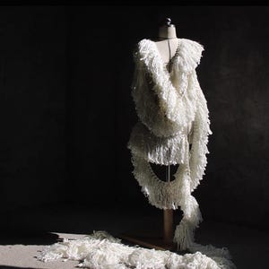 May include: A white, fringed garment on a mannequin. The dress-like design has long, fringed elements. The piece is set against a dark background, highlighting its intricate details. The garment appears to be made of a soft material.