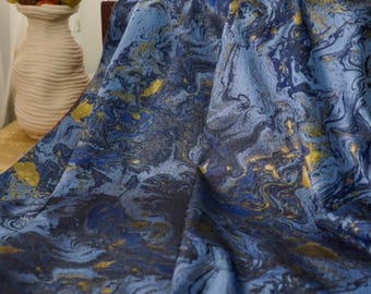 May include: A blue fabric with a marbled design, featuring shades of navy, light blue, and gold. The fabric is draped, showcasing its texture and pattern. The overall theme is textile and home decor.