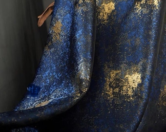 May include: Close-up of a draped fabric with a dark blue base and gold metallic accents. The fabric has a textured appearance, with the gold highlights creating a mottled effect across the surface. The folds of the fabric are visible.