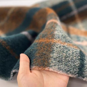 Puede incluir: Primer plano de una tela doblada con un patrón de cuadros. La tela presenta una combinación de colores verde azulado, marrón y naranja. La textura parece suave y difusa, con un tejido visible. El diseño de cuadros está compuesto por rayas que se cruzan.