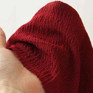 Pode incluir: Close-up de um tecido vermelho escuro com uma superfície texturizada e enrugada. O tecido é segurado, mostrando suas dobras e o jogo de luz e sombra em sua superfície. O material parece macio e maleável.