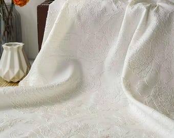 May include: Off-white fabric with a floral pattern. The fabric is draped, showing the texture and design. A vase with flowers and a decorative vase are in the background.