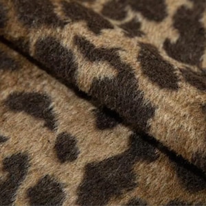 Könnte beinhalten: Nahaufnahme eines Stoffes mit Leopardenmuster. Der Stoff hat einen beigefarbenen Hintergrund mit dunkelbraunen Flecken. Die Textur ist weich und leicht flauschig, mit sichtbaren Falten und Schatten.