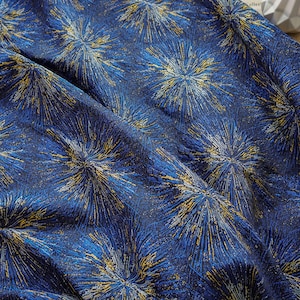 May include: Close-up of a textured fabric with a repeating pattern of starburst-like designs. The fabric is primarily a deep blue, with accents of gold, light blue, and white. The folds in the fabric create shadows and depth.