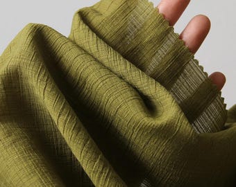 May include: Close-up of olive green fabric with a textured, pleated design. The fabric is held up, showing the weave and the way it drapes. The color is a muted, earthy tone, and the fabric appears lightweight.