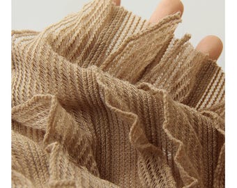 May include: Close-up of a beige, open-weave fabric with a ruffled edge. The fabric has a textured appearance and is held in place by fingers. The material appears to be lightweight and airy, suitable for clothing or accessories.