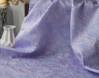 May include: A draped, lavender fabric with a raised rose pattern. The fabric is folded, creating shadows and highlights that emphasize the texture. The background includes a vase with flowers.