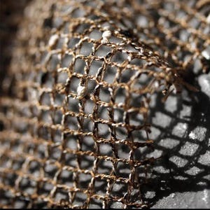 May include: Close-up of a woven net with a grid-like pattern. The net is made of brown and white threads, creating a textured surface. The image is well-lit, highlighting the details of the weave and the shadows.