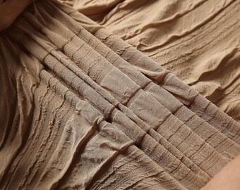 May include: Close-up of a beige fabric with a textured, pleated design. The fabric has a subtle sheen and is gathered in folds, creating a layered effect. The material appears soft and flowing, with a detailed pattern.