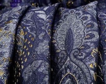 May include: Close-up of a navy blue fabric with a paisley pattern in silver and gold. The fabric is folded, creating deep shadows and highlighting the texture and sheen of the material. The fabric has a luxurious appearance.