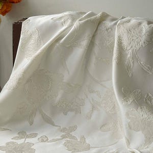 May include: Cream-coloured fabric with a floral pattern. The fabric is draped, showing the texture and design of the raised floral elements. The flowers and leaves are in a tone-on-tone design.