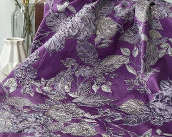 May include: A close-up of a purple fabric with silver floral embroidery. The fabric is draped, showcasing the intricate details of the embroidered flowers and leaves. The color palette includes shades of purple and silver.