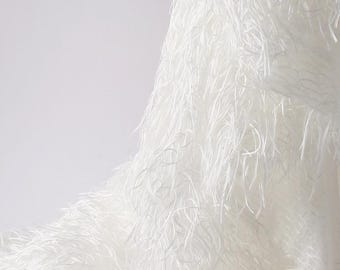 May include: Close-up of a white fabric with a textured surface and long, thin strands. The material has a soft, feathery appearance, creating a flowing effect. The fabric is likely used for clothing or decorative items.