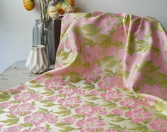 May include: A floral fabric with a repeating pattern of pink flowers and green leaves on a cream background. The fabric is draped over a surface, showcasing its texture and design. The fabric is likely used for home decor or crafting.