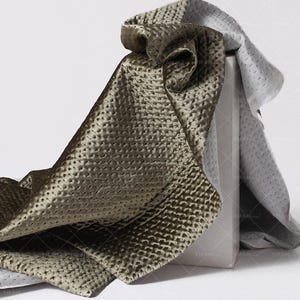 May include: Olive-green textured fabric with a quilted pattern, draped over a white box. The fabric reveals a lighter grey fabric with a similar pattern. The image suggests a luxurious, tactile textile with a subtle sheen.