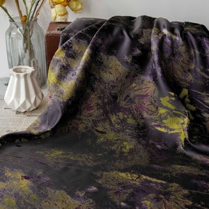 May include: A draped fabric with a dark purple base and a pattern of yellow and purple brushstrokes. The fabric is on a table with a vase of flowers and a white geometric vase.