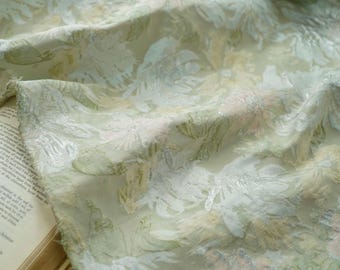 May include: Close-up of a delicate fabric with a floral pattern. The fabric features a soft, flowing texture with a light green, white, and pale pink color palette. The floral design is intricately detailed, creating a sense of elegance.