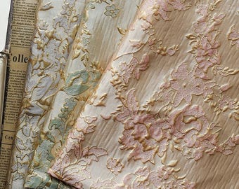 May include: Close-up of several folded fabrics with floral patterns. The fabrics feature a cream-colored base with embroidered designs in gold, pink, and light green. The floral patterns include roses and other stylized flowers.