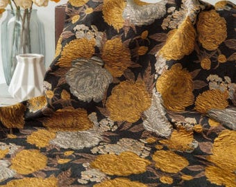 May include: A close-up of a black fabric with a floral pattern. The design features large, textured flowers in shades of gold and silver, with brown leaves. The fabric is draped, creating shadows and depth. The overall effect is rich and luxurious.