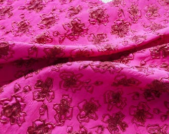 May include: Bright pink fabric with a floral pattern in a darker shade of pink. The fabric is textured and appears to be draped, showcasing the intricate floral design. Ideal for apparel or home decor projects.