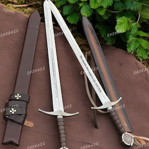 May include: Two silver-toned steel swords with brown leather-wrapped hilts and circular pommels featuring red accents. One blade is engraved with the text "The battle belongs to the LORD" and the other with Latin text. Includes two brown leather scabbards.